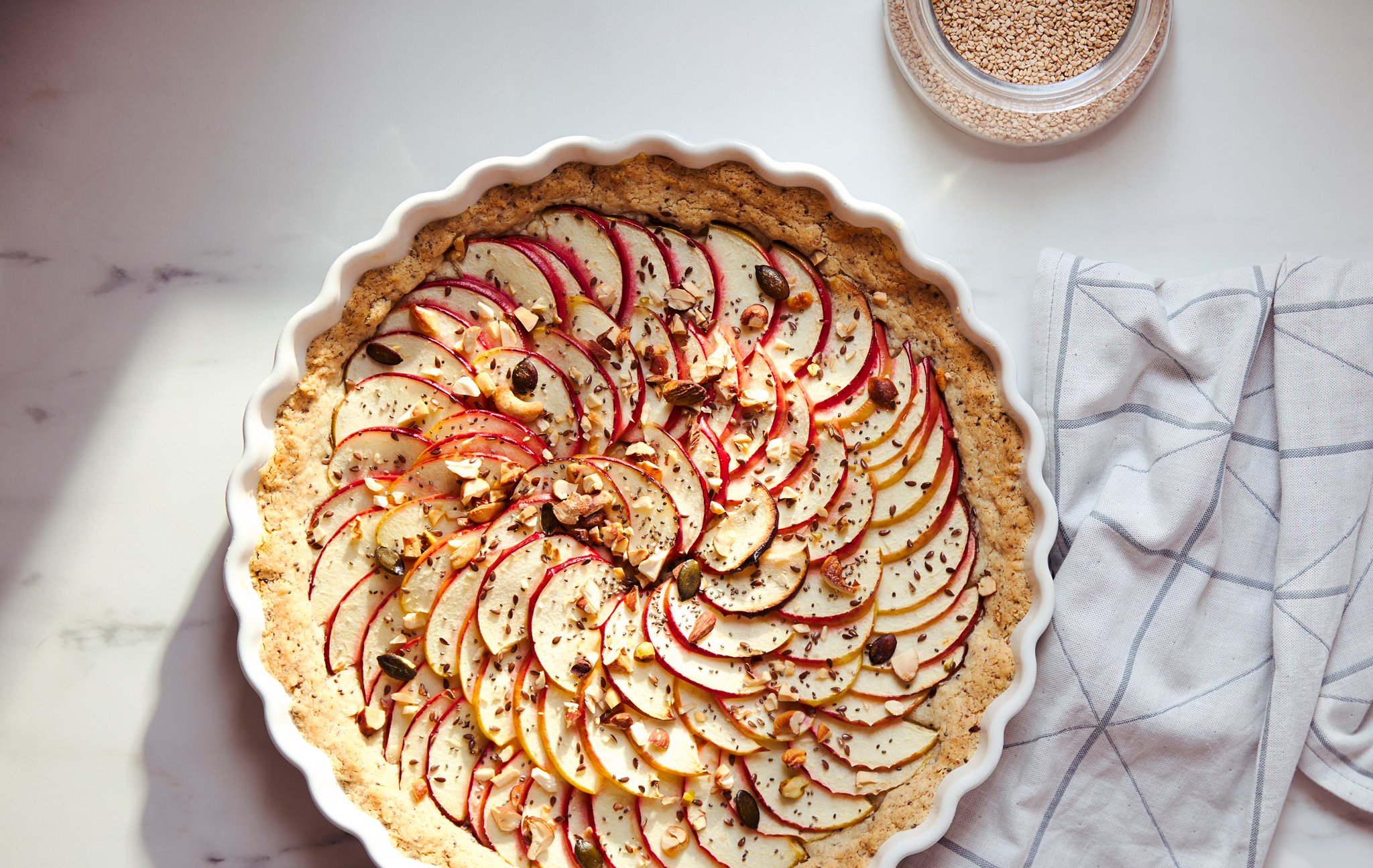 A white kitchen towel with a grey, graphic pattern and a pie covered with thin apple slices in a white VARDAGEN pie dish.