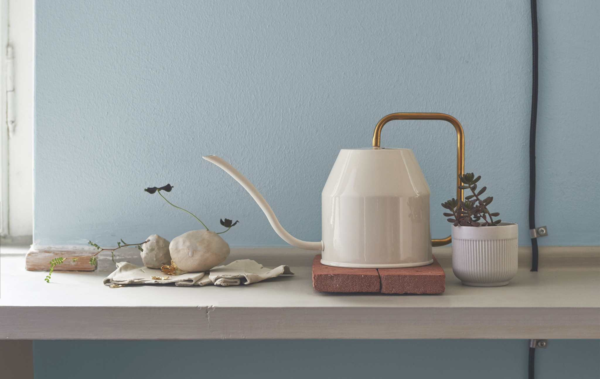 A white watering can with gold-colour handle on a shelf with pot plant and stones.