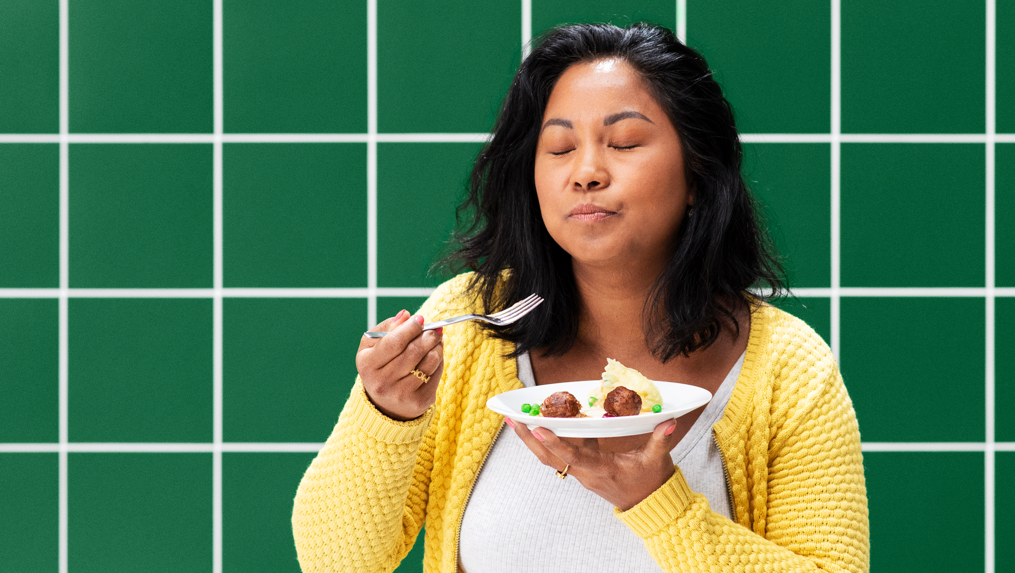A woman closing her eyes while holding a plate, tasting plant balls with mashed potatoes and green peas.