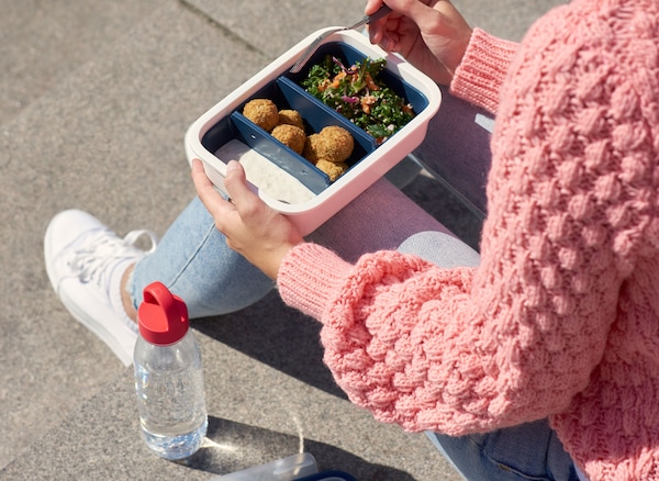 A woman eating from an IKEA plastic food container, with a transparent plastic bottle filled with water beside her.