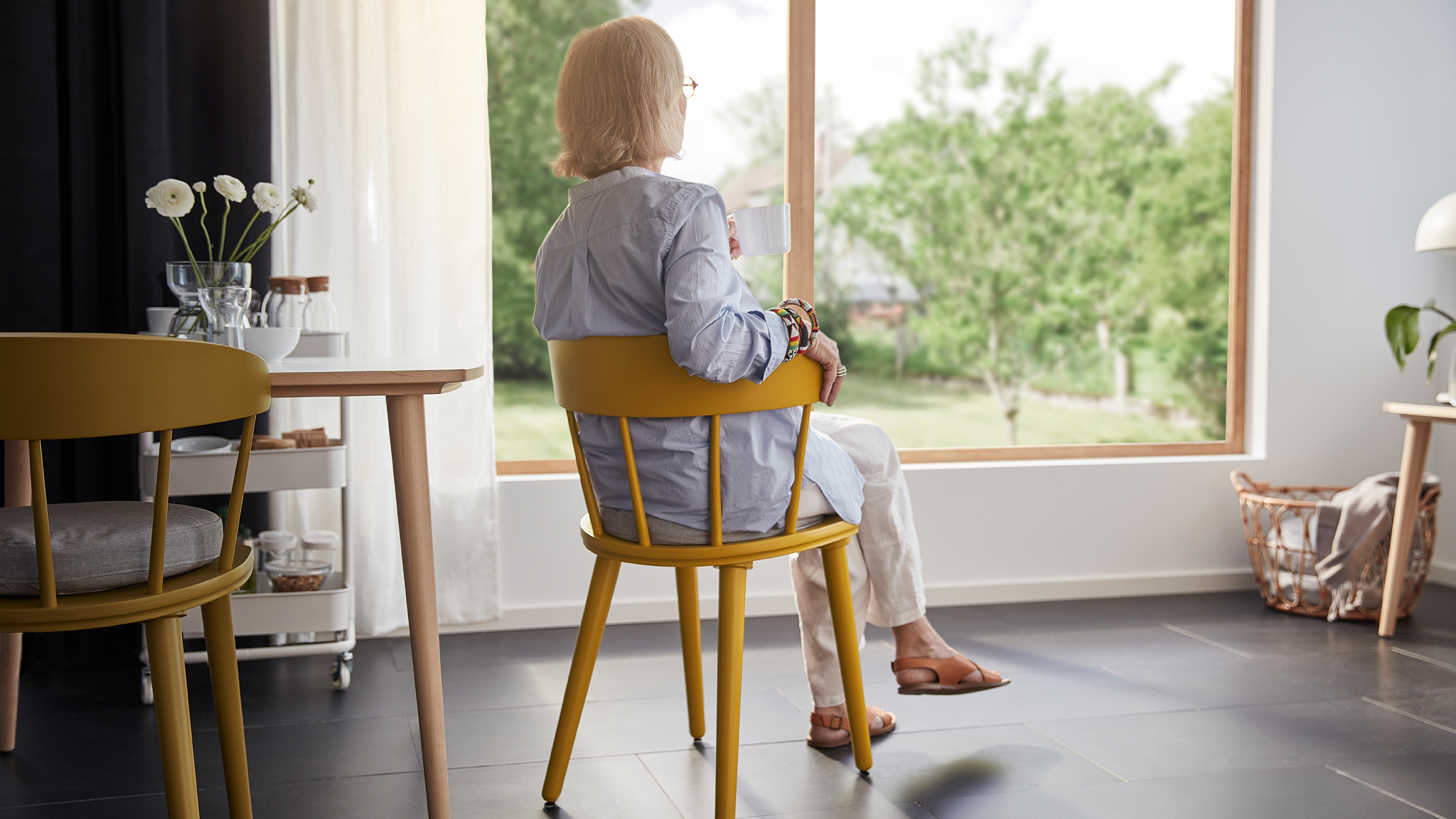 A woman in her 60s sits comfortably in the OMTÄNKSAM chair. She has her back to us and looks out the living room window.
