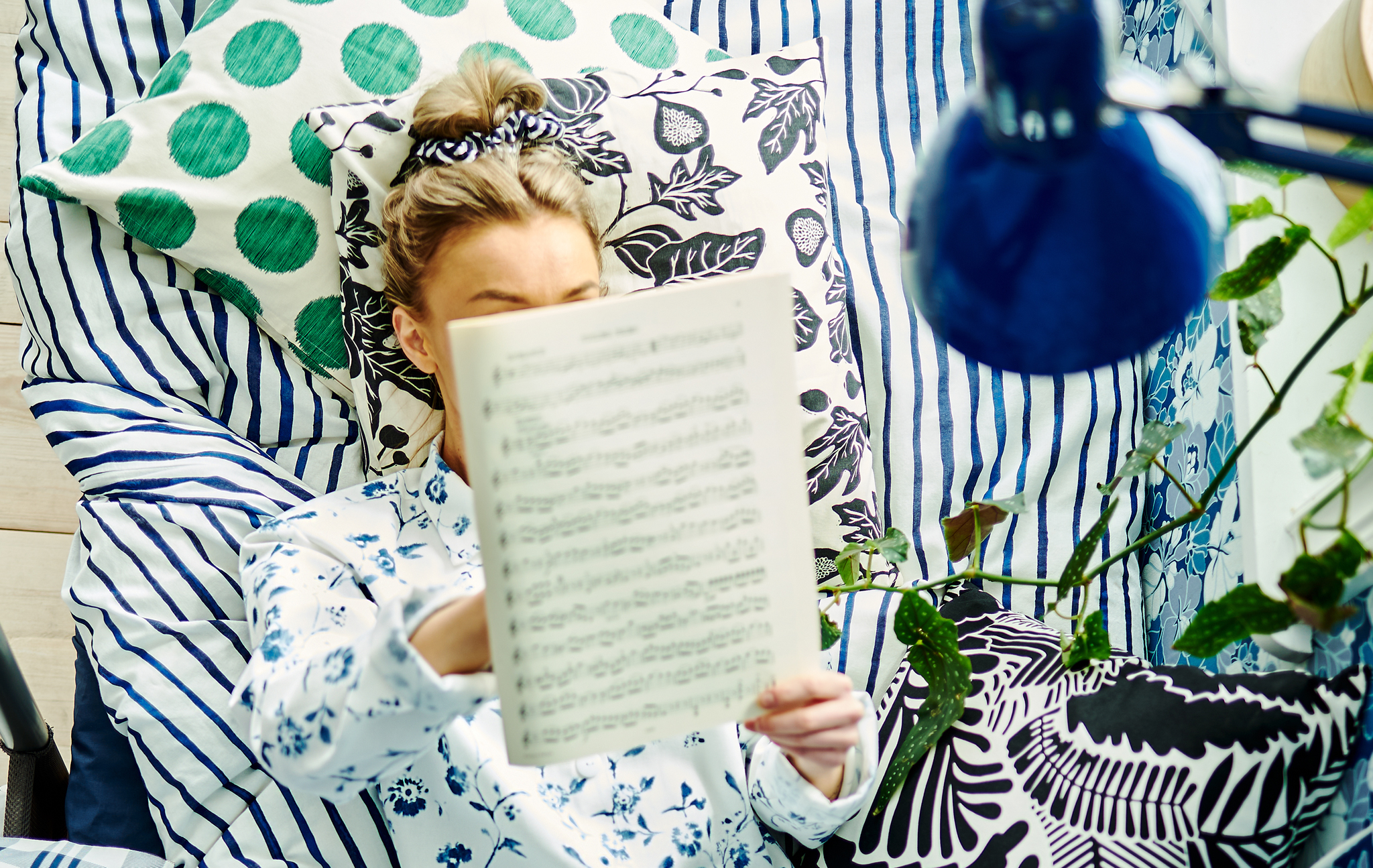 A woman lying in bed with cushions, bed linen and wallpaper with different patterns around her, while reading musical notes.
