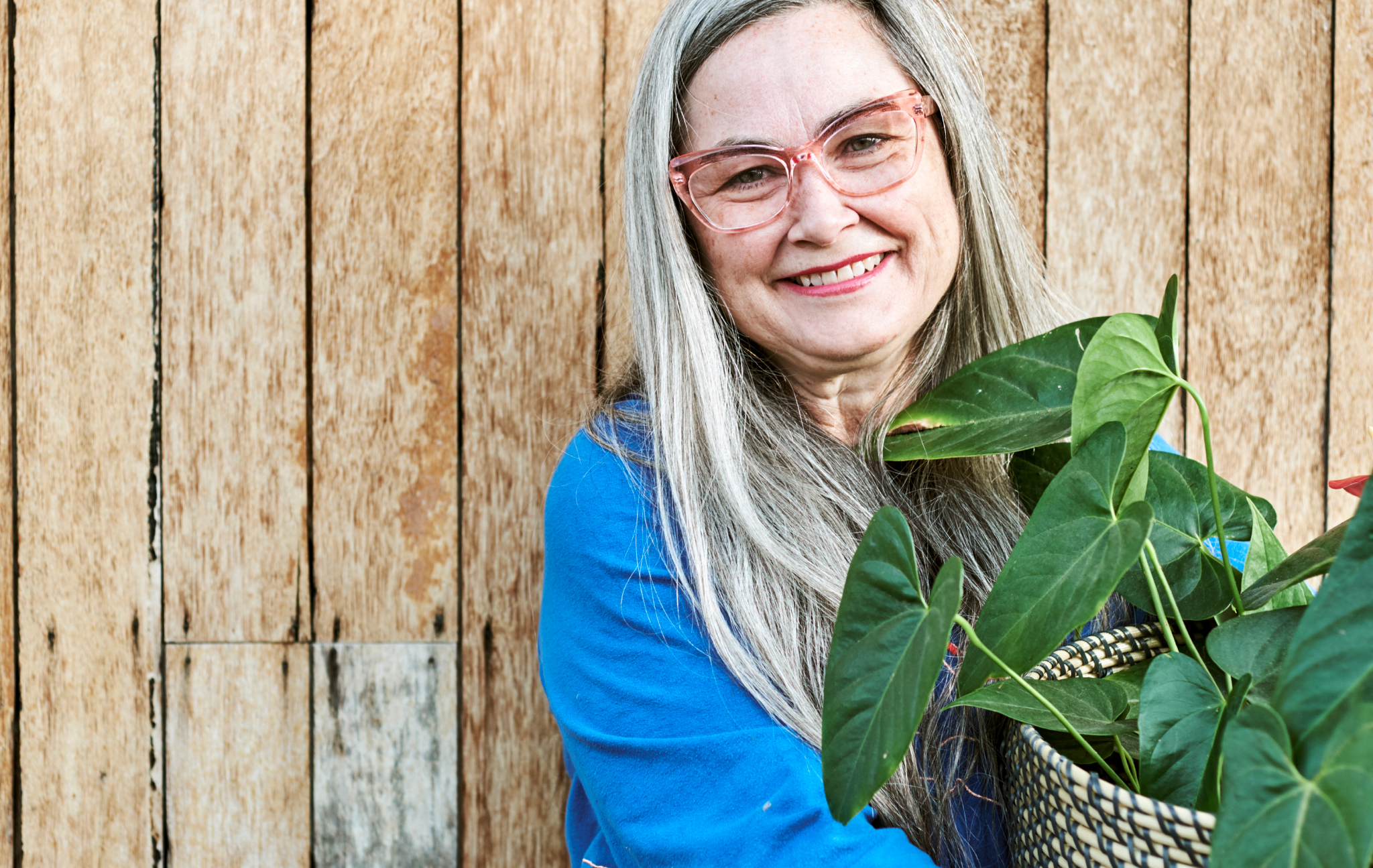A woman with long grey hair, wearing pink-rimmed glasses and blue top, smiles and holds a leafy plant in a woven plant pot.