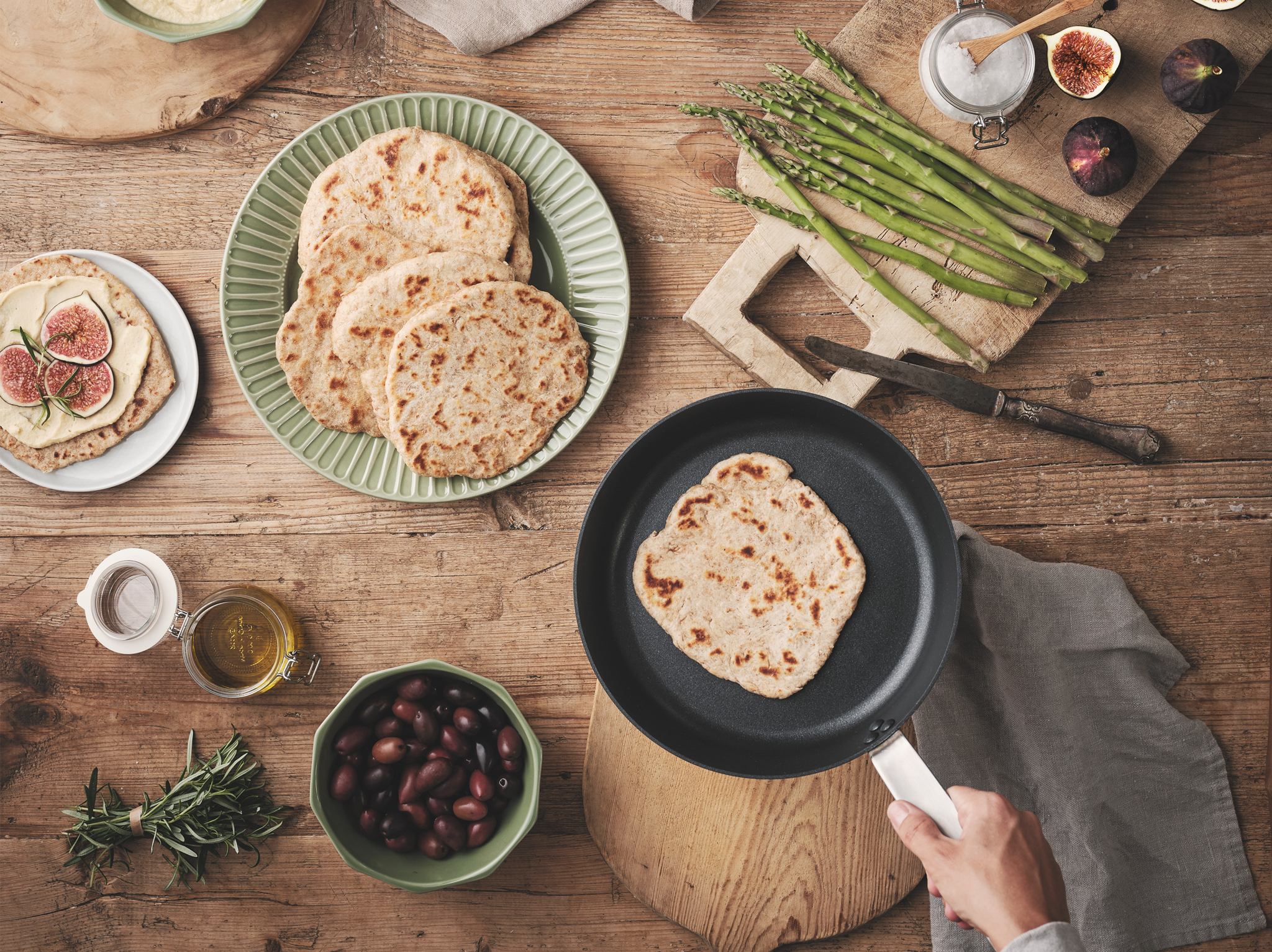 A wooden table with KNÅDA flat bread on a green plate and a frying pan, with condiments around it like asparagus and olives.
