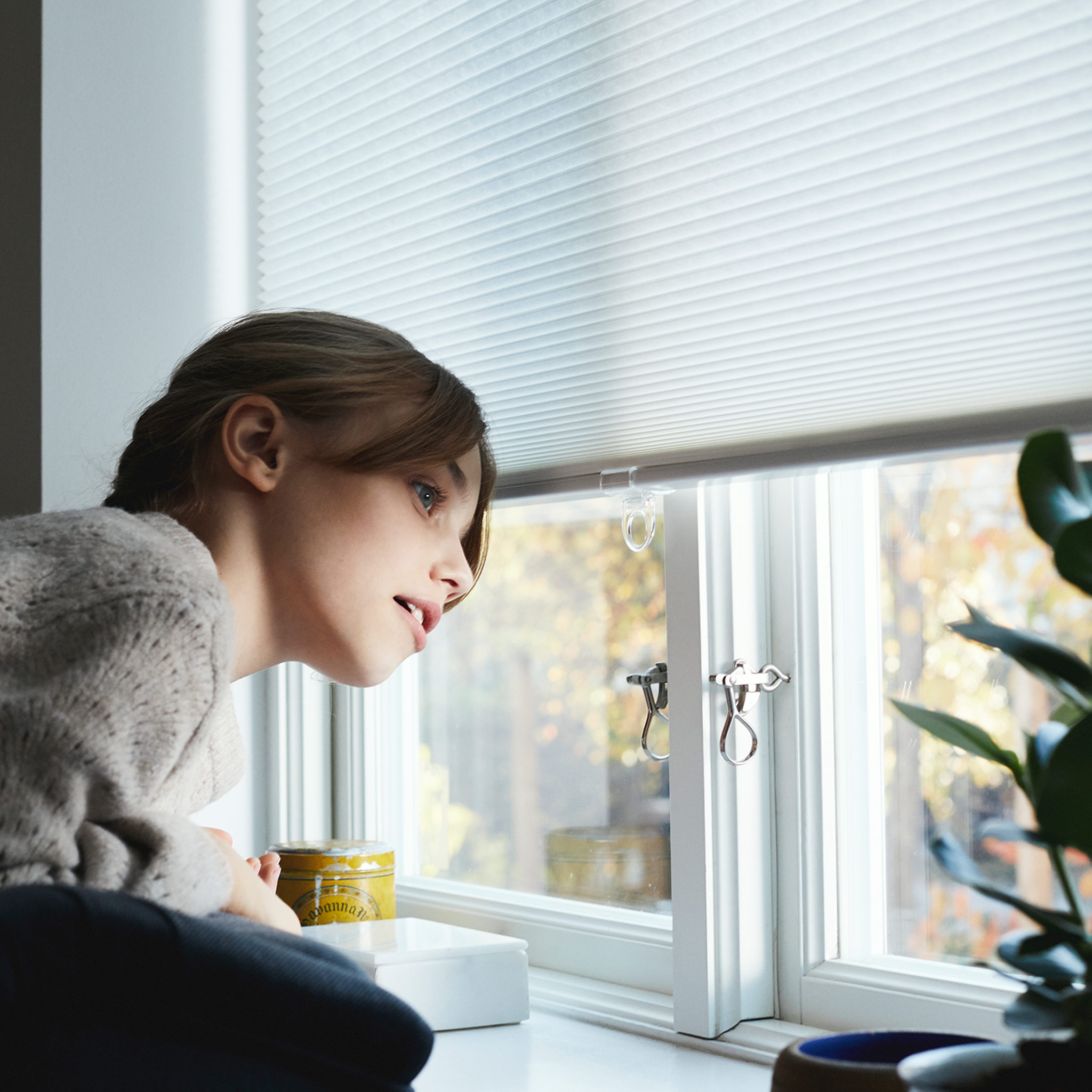 A young girl wearing a sweaters looks out the window underneath the blinds.
