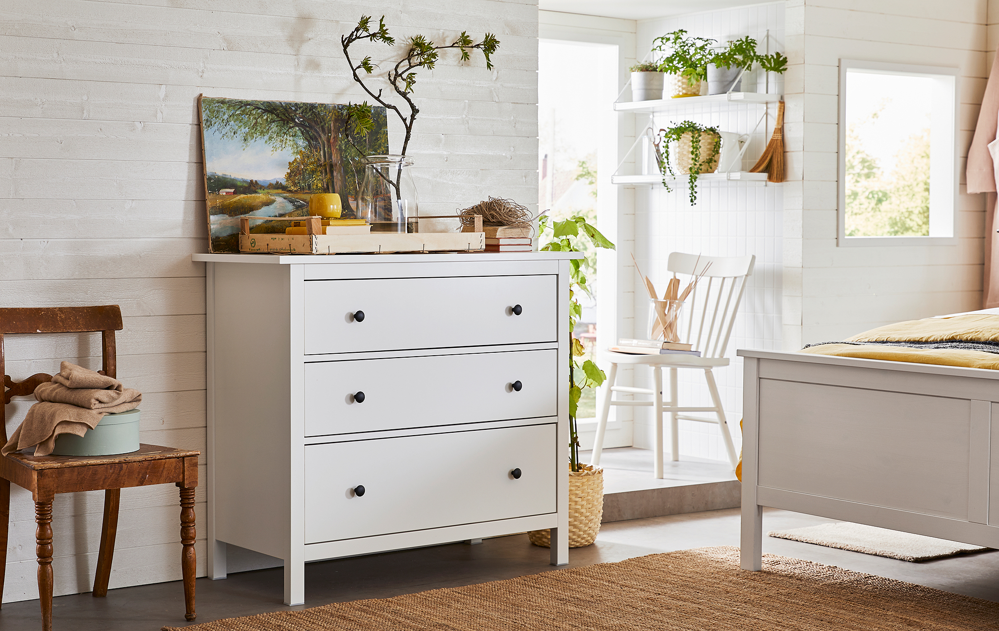 Airy bedroom interior dominated by wood choices and greenery, a three-drawer chest standing close to the bed’s end.