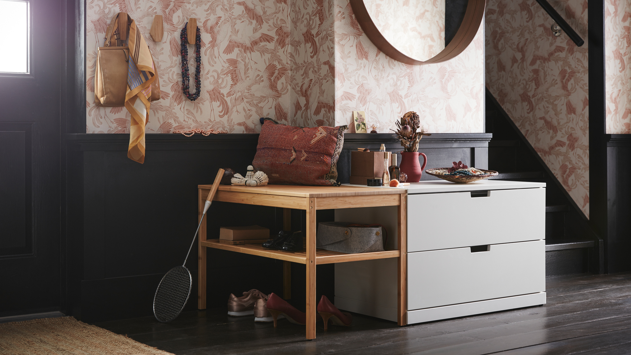An entrance with a light grey NORDLI chest of two drawers under a round mirror and beside a wooden bench with open shelves.
