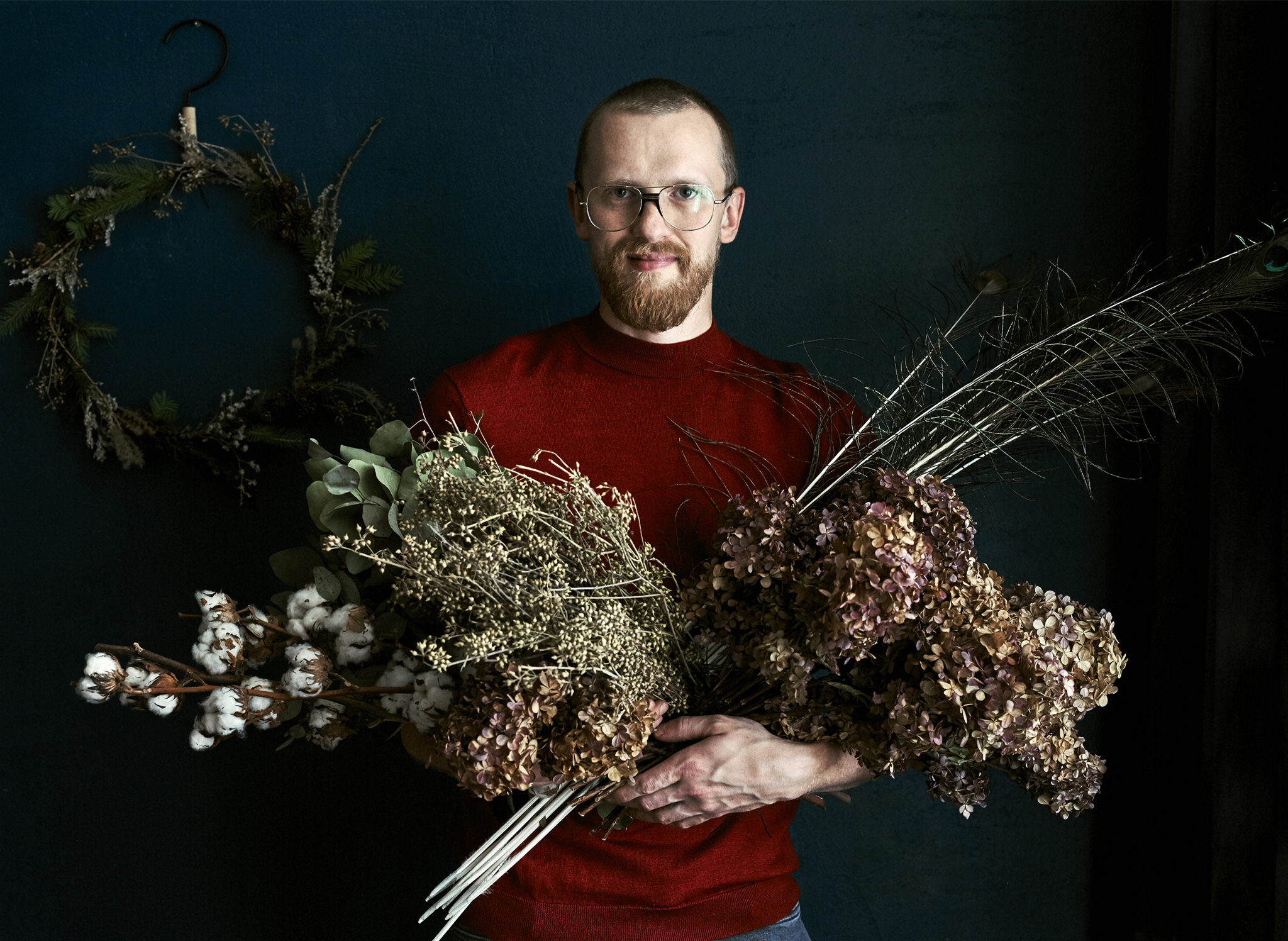 An image of Łukasz standing in front of a wreath holding armfuls of dried foliage.
