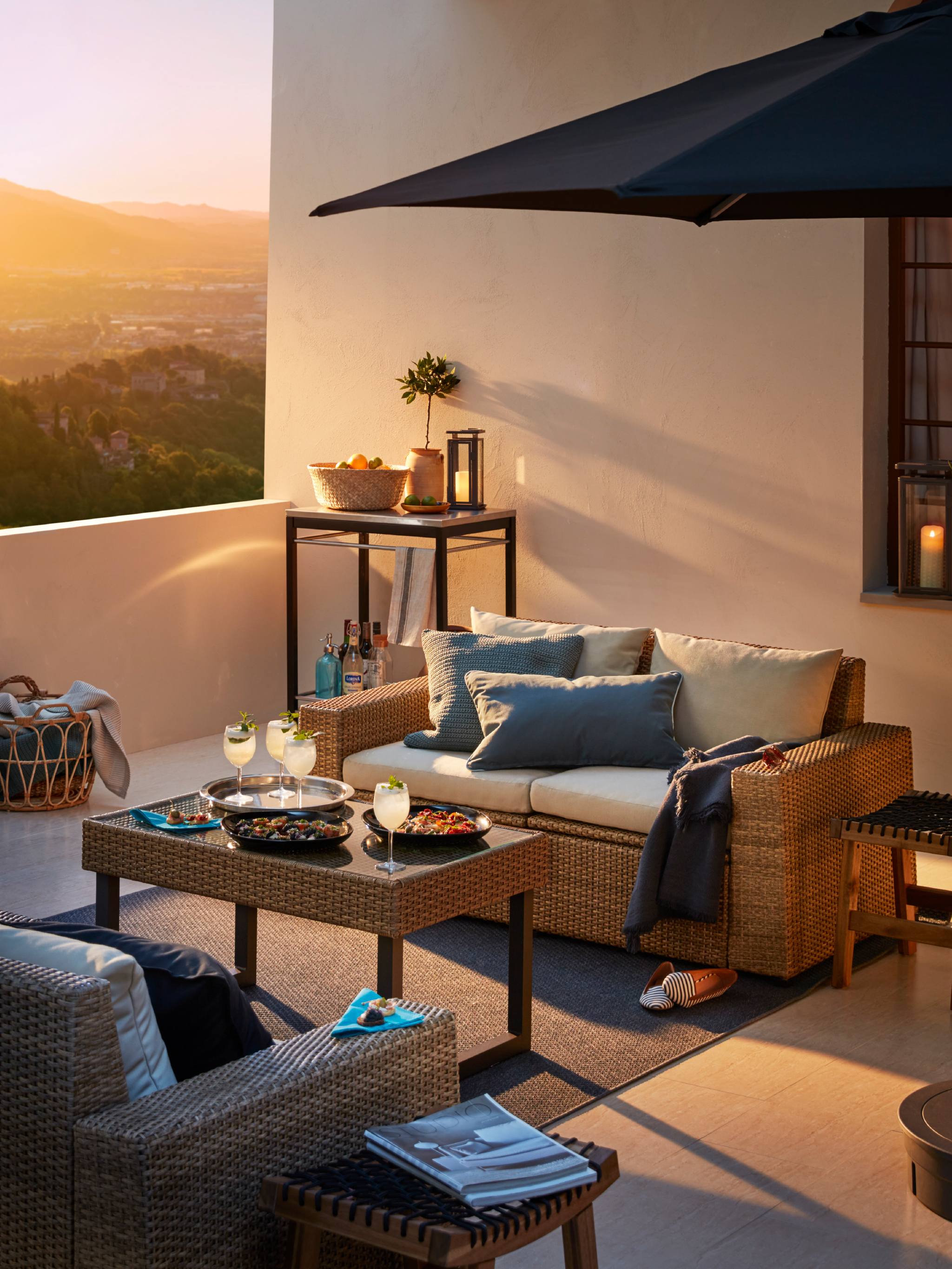 An outdoor sofa with white cushions on a terrace with a parasol, coffee table with refreshments and an outdoor armchair.