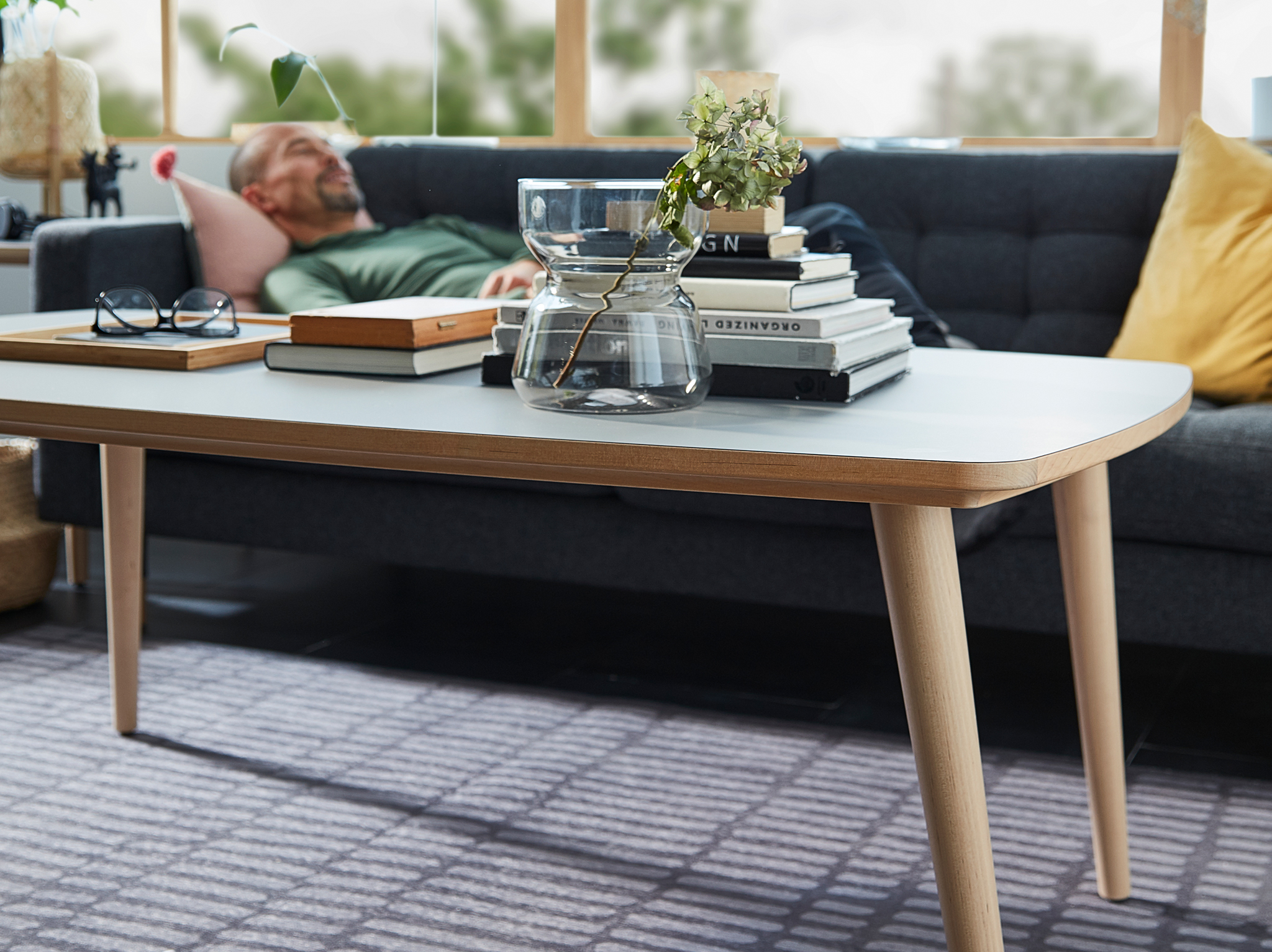 OMTÄNKSAM coffee table, with OMTÄNKSAM vase, books and glasses on top, is in focus. A man rests on a sofa in the background.