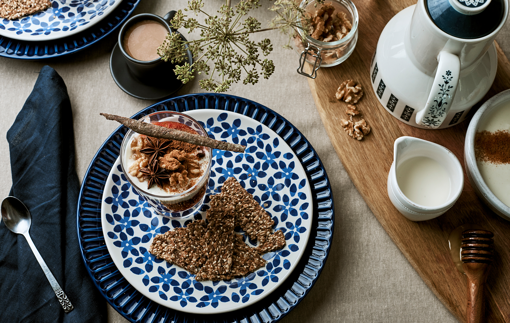Breakfast table set with linen, patterned blue plates, granola in a glass and coffee accessories on a chopping board.