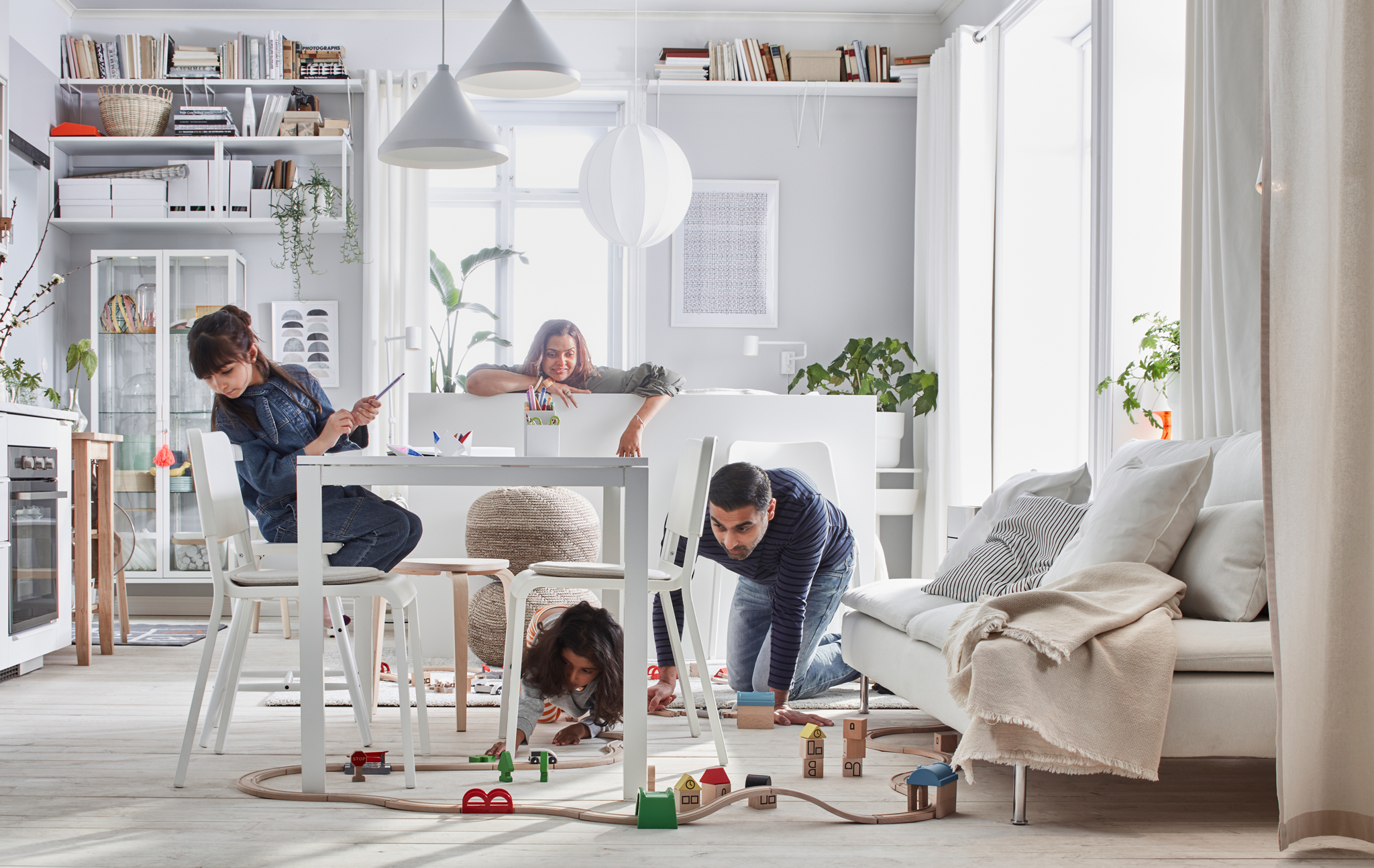 Compact-living home where a family plays with a LILLABO train set in a room with bed, sofa, table, kitchenette, and storage.