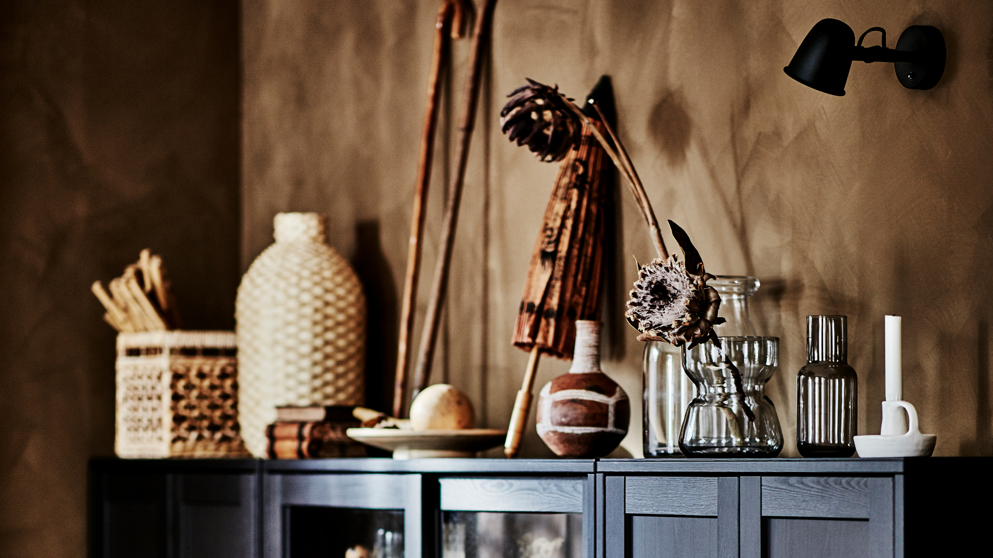 Corner of a living room with a dark, wide HAVSTA cabinet filled and covered with artful objects in natural materials.