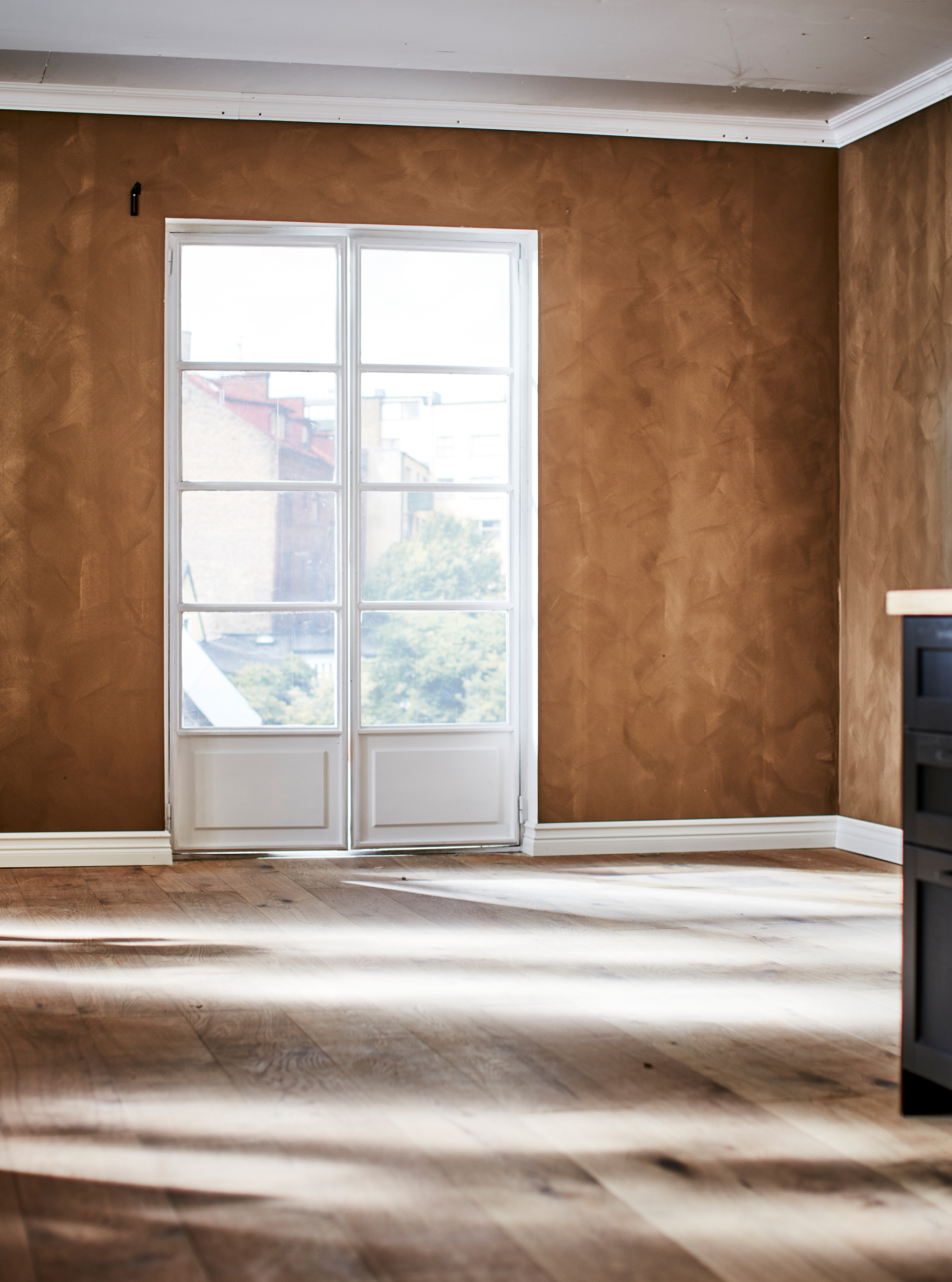 Empty room with French windows, a wooden floor, and the wall painted in a rich, brown tone with a matte suede finish.