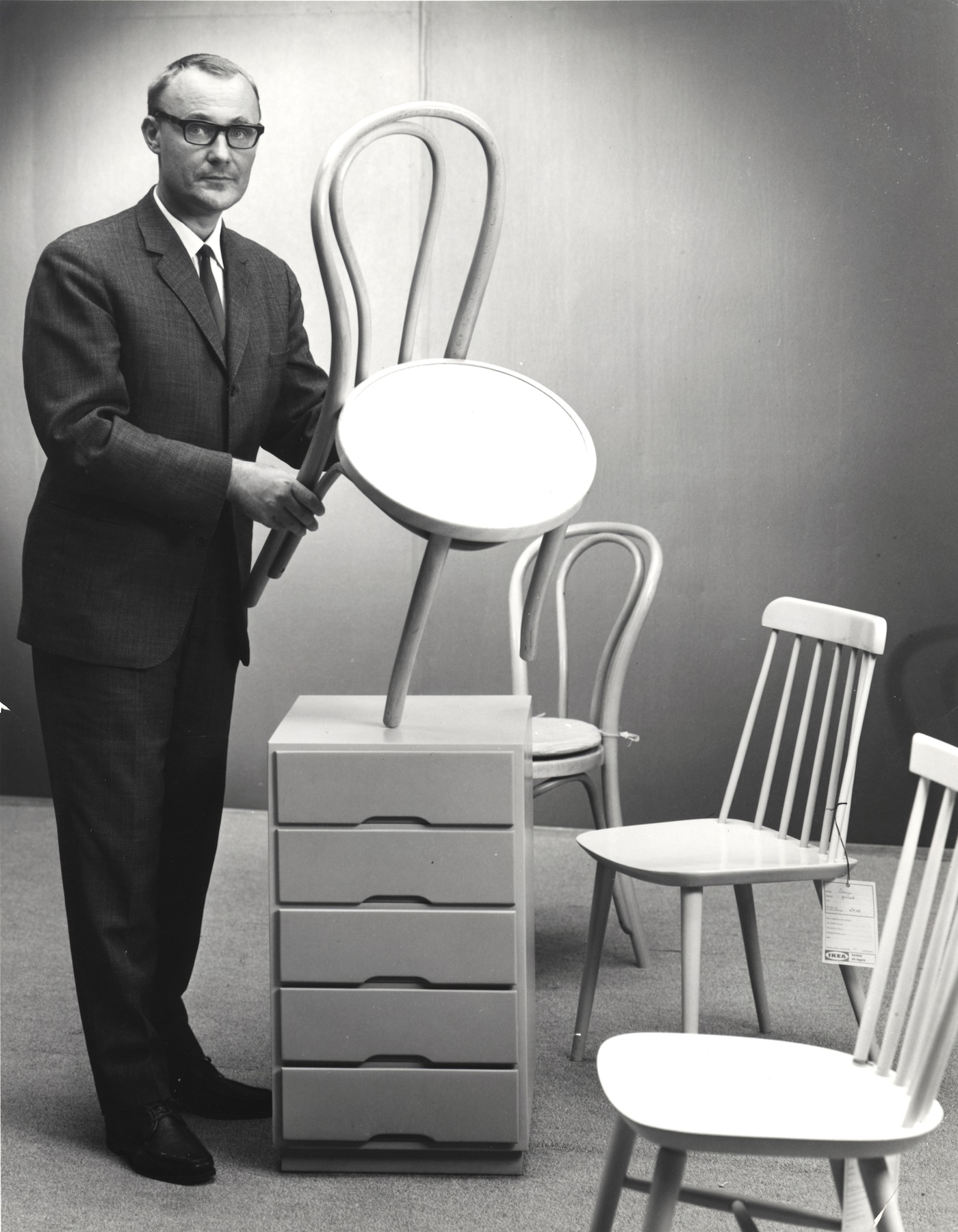 A black and white photo of a young Ingvar Kamprad standing in a studio holding an IKEA chair. 