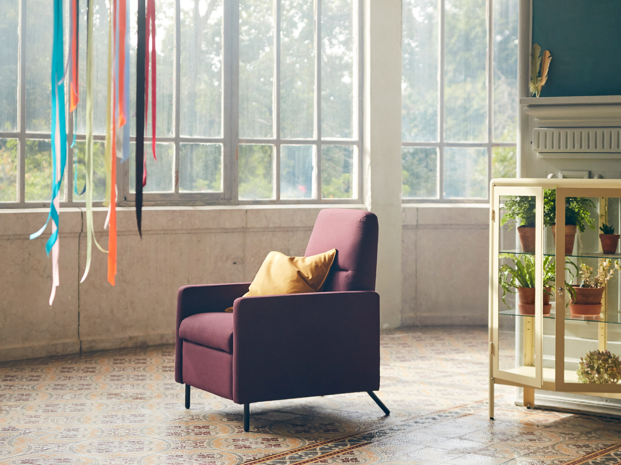 GISTAD recliner in a dark red colour with a yellow cushion placed in its seat, standing in a room with big windows.