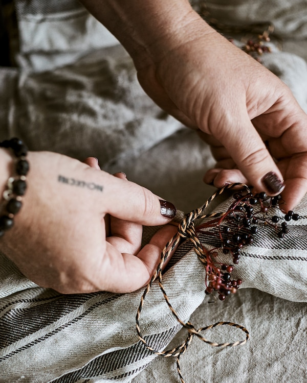 Hands tying a decorative knot made from natural and black coloured twine and plum coloured wild berries round a napkin.