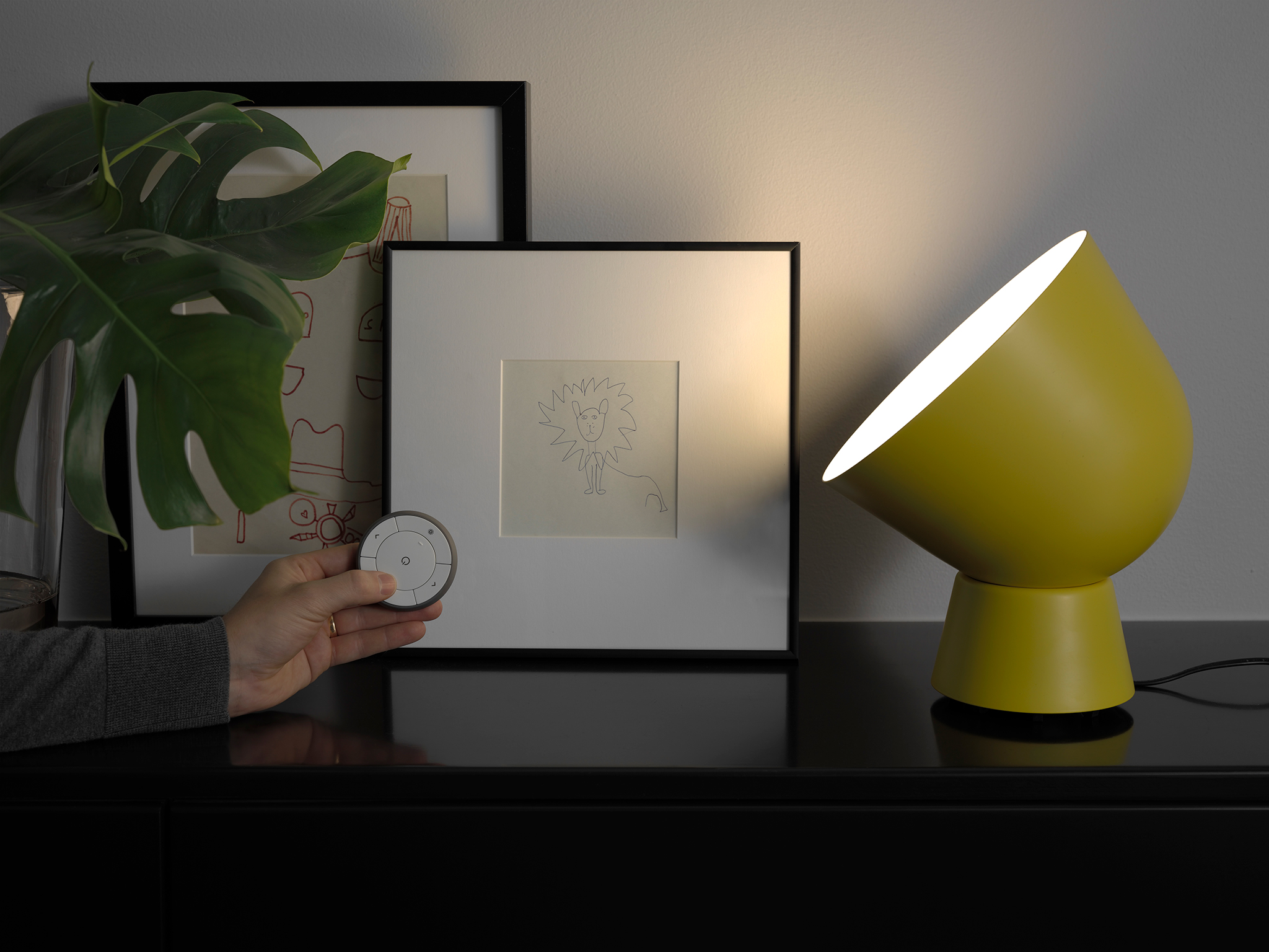 IKEA PS 2017 table lamp in front of two picture frames and a plant, with a hand holding a small circular watch-like object.
