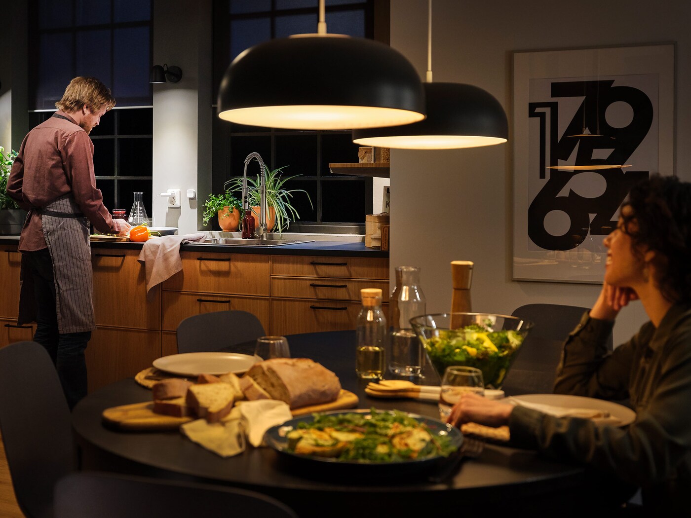 Kitchen with large pendant lamps with smart lighting and a table set for dinner, a woman sitting at the table and a man at the sink.