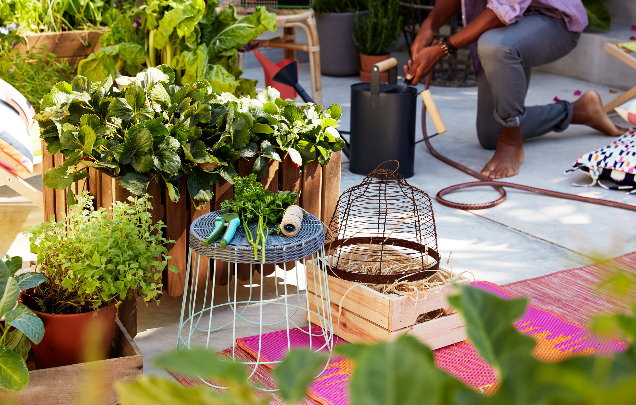 Large wooden plant crate with plants standing on concrete outdoors, surrounded by rugs, a stool, garden tools.