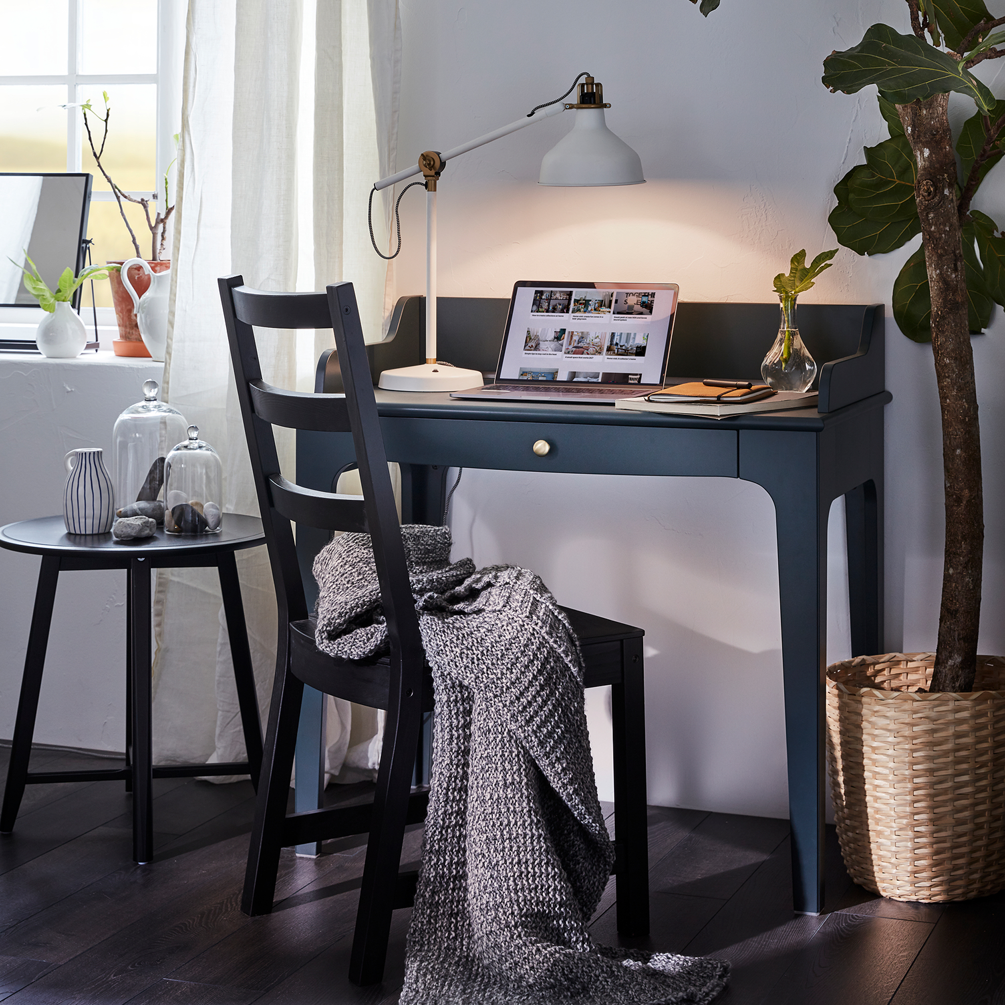 LOMMARP dark blue-green desk has a laptop and a lit table lamp on it and in front of it is a black chair with a grey throw.