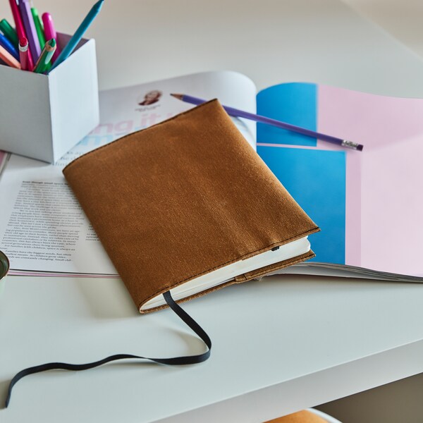 Notebook protected by LANKMOJ notebook cover in brown, placed on a workspace table with other note books, pens and pencils.