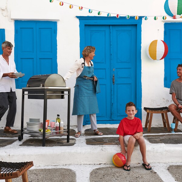 Outdoor scene, with a man and woman standing by a grill with a boy sitting on the floor.