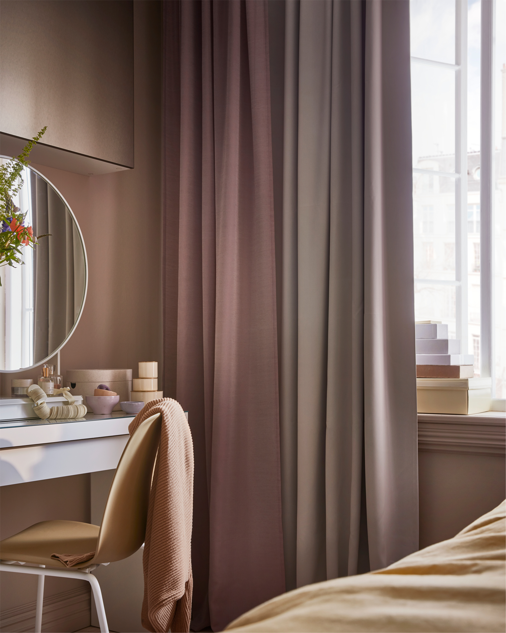 Pink and grey curtains, a yellow chair, and white dressing table with a white table mirror and small boxes standing on it.