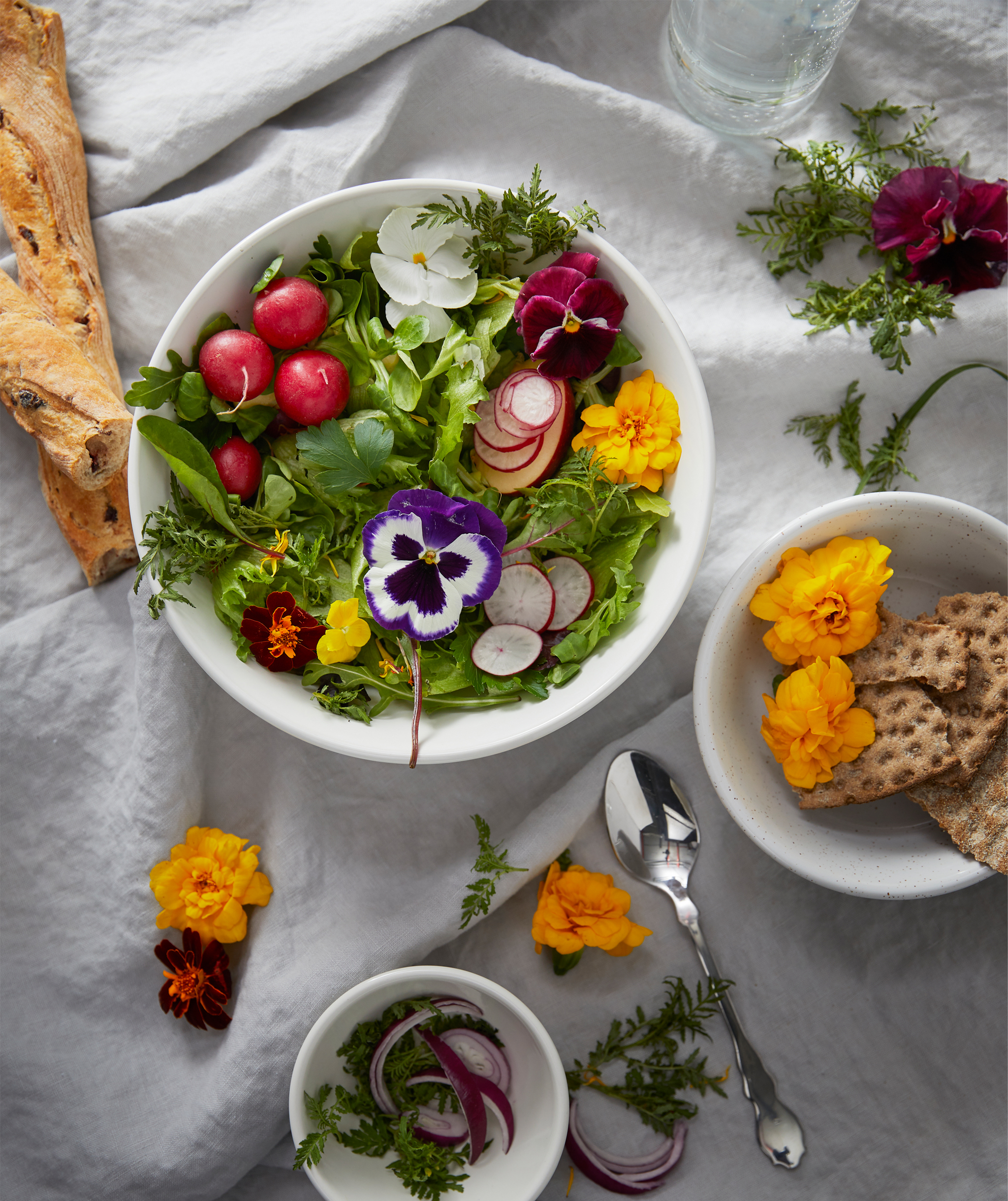 Section of a table set with a white, creased cloth, different-size bowl filled with bread, homegrown greens, and flowers.