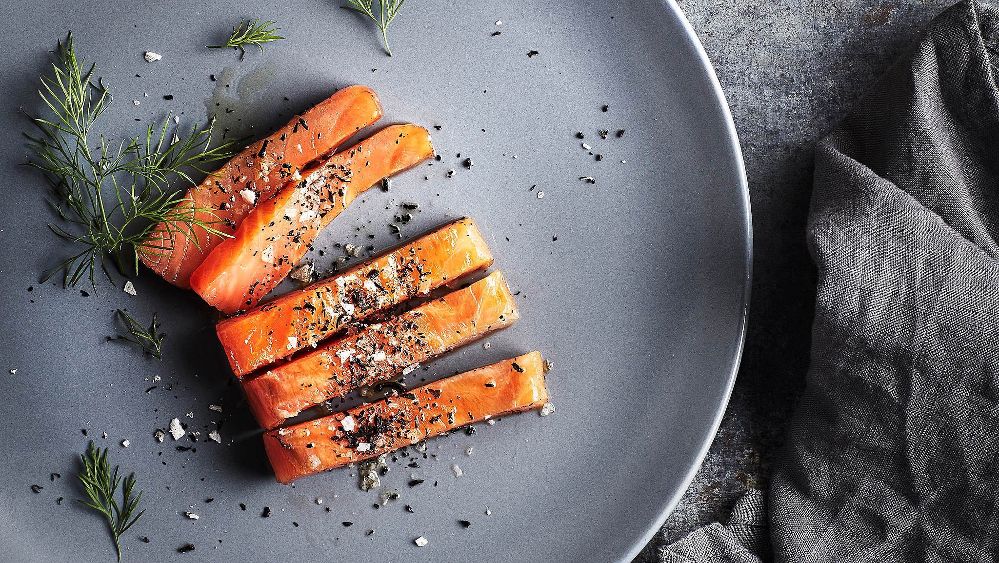Slices of salmon covered with a sprinkling of salt, pepper and dill on a grey plate. A grey napkin is beside the plate.