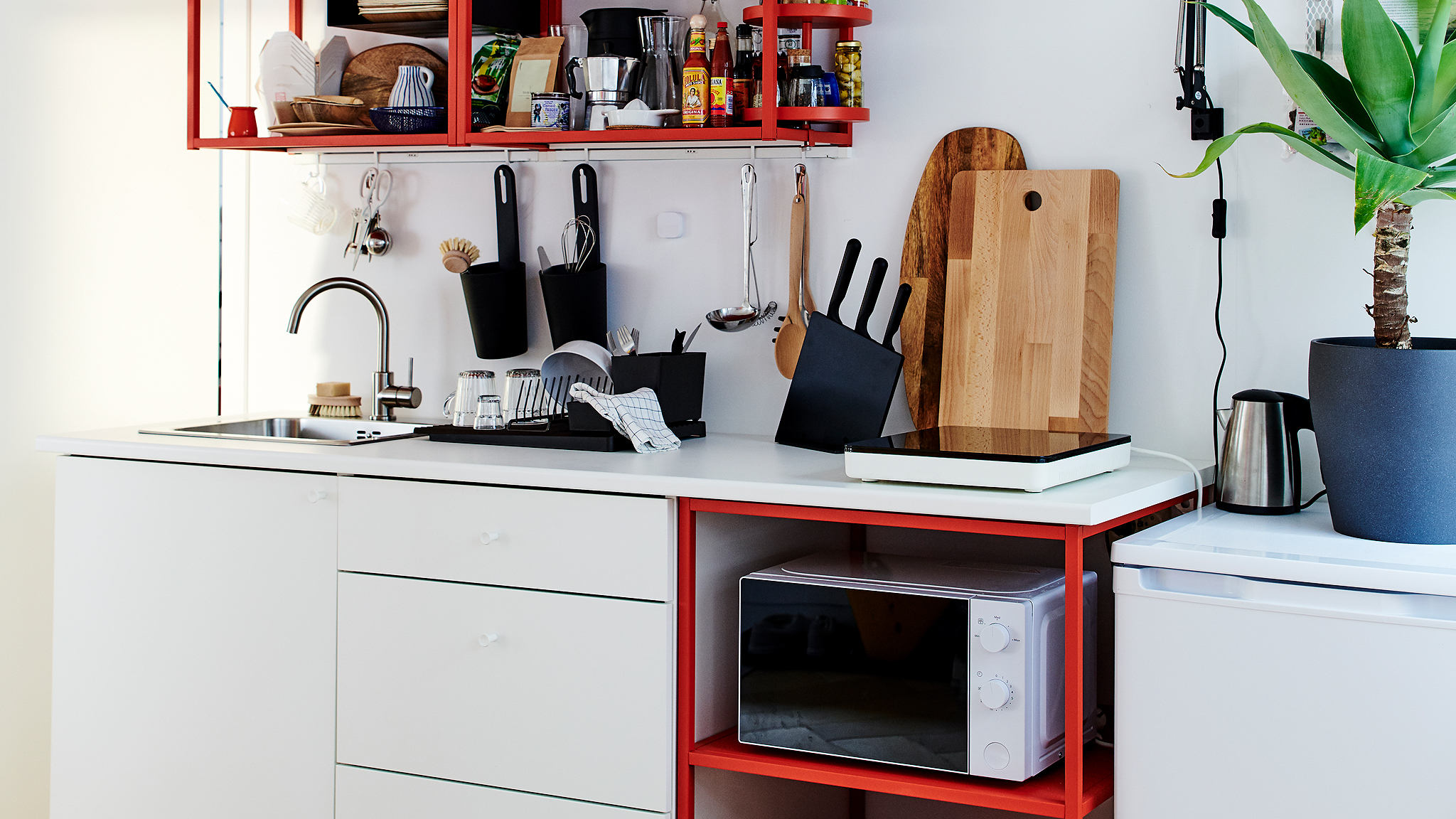 Small kitchen in red/white with a portable induction hob, wooden chopping boards, and a black dish drainer.