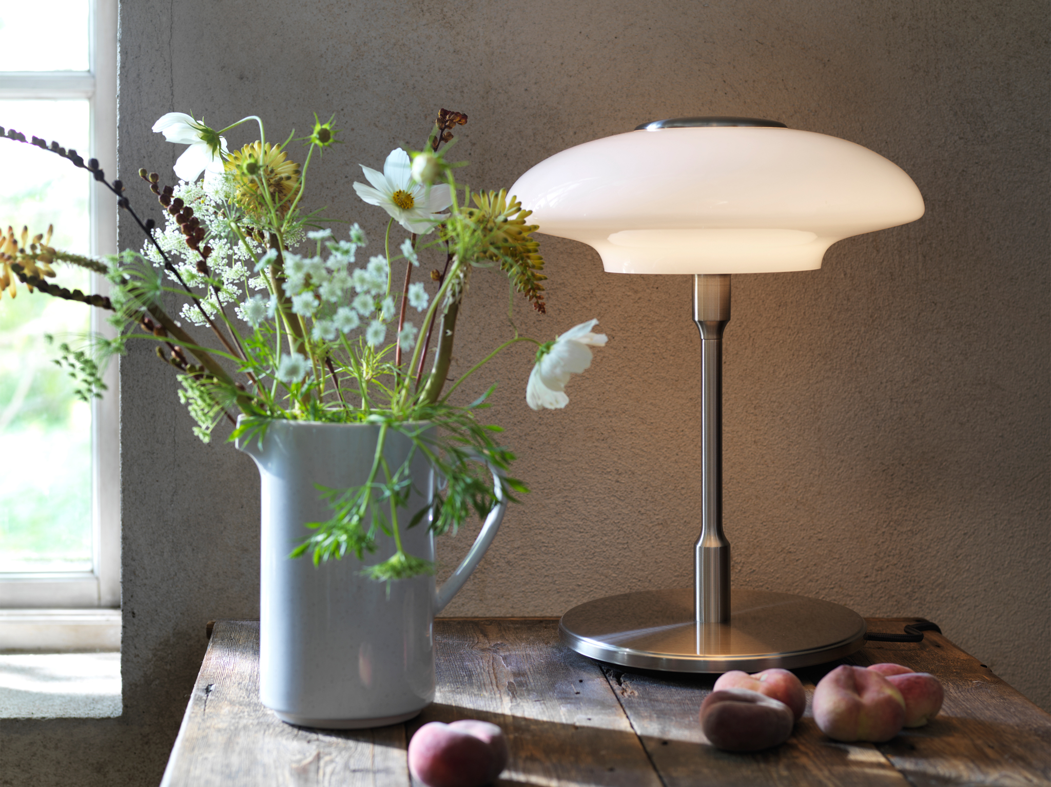 TÄLLBYN table lamp designed with Art Deco inspiration placed on a wooden table with a jug of flowers and peaches.
