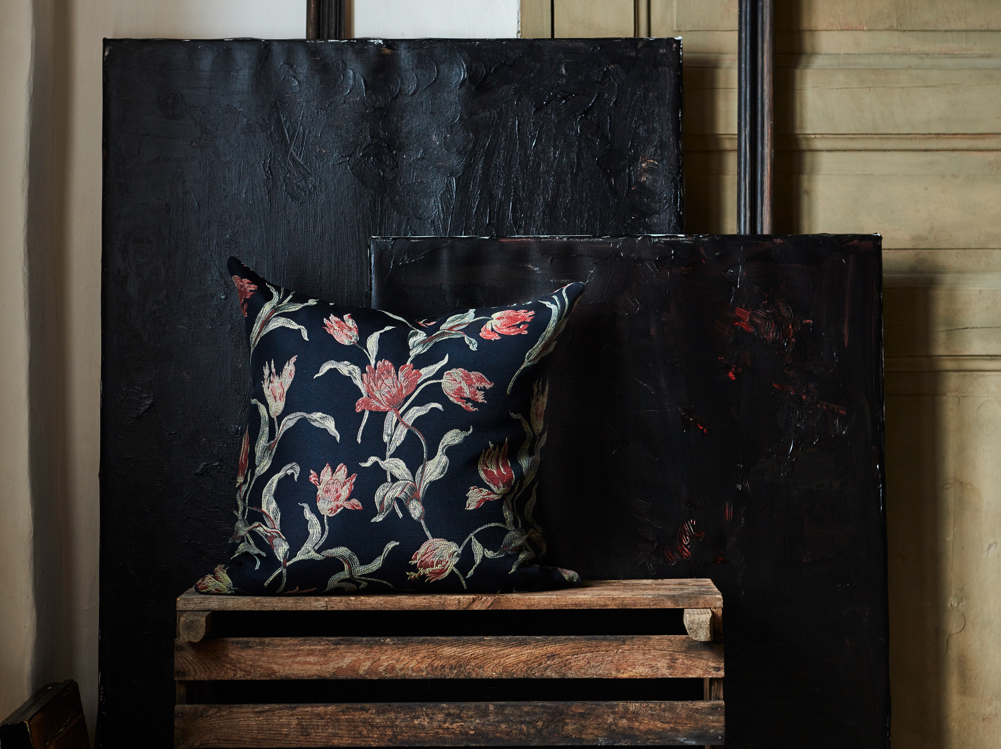 The hand-embroidered ÅLANDSROT cushion with a tulip pattern on a dark-blue background placed on wooden crates in a dark room.