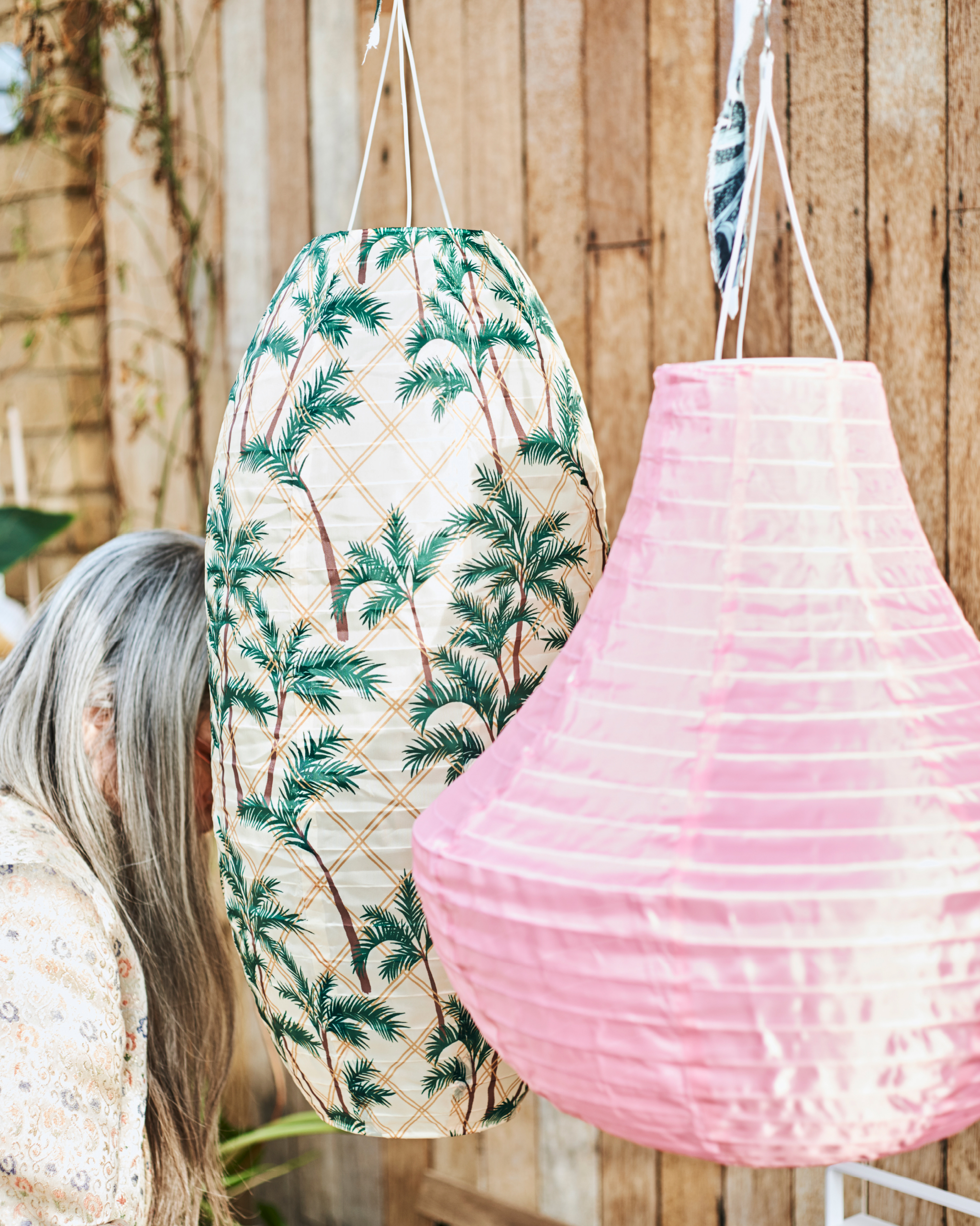 Two big hanging outdoor lanterns, one in pink, the other with palm tree print, partially hiding a woman with long grey hair.