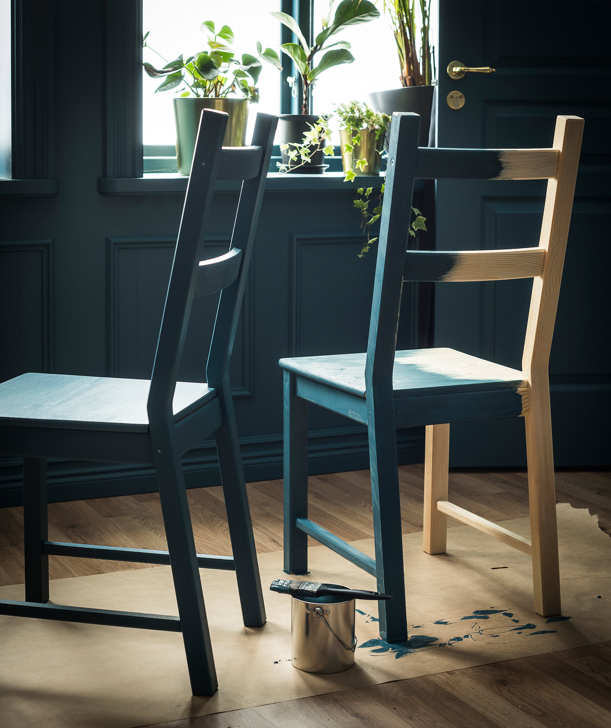 Two blond-wood chairs placed on floor-protective paper. Otherwise untreated, the chairs are being painted in a dark shade.