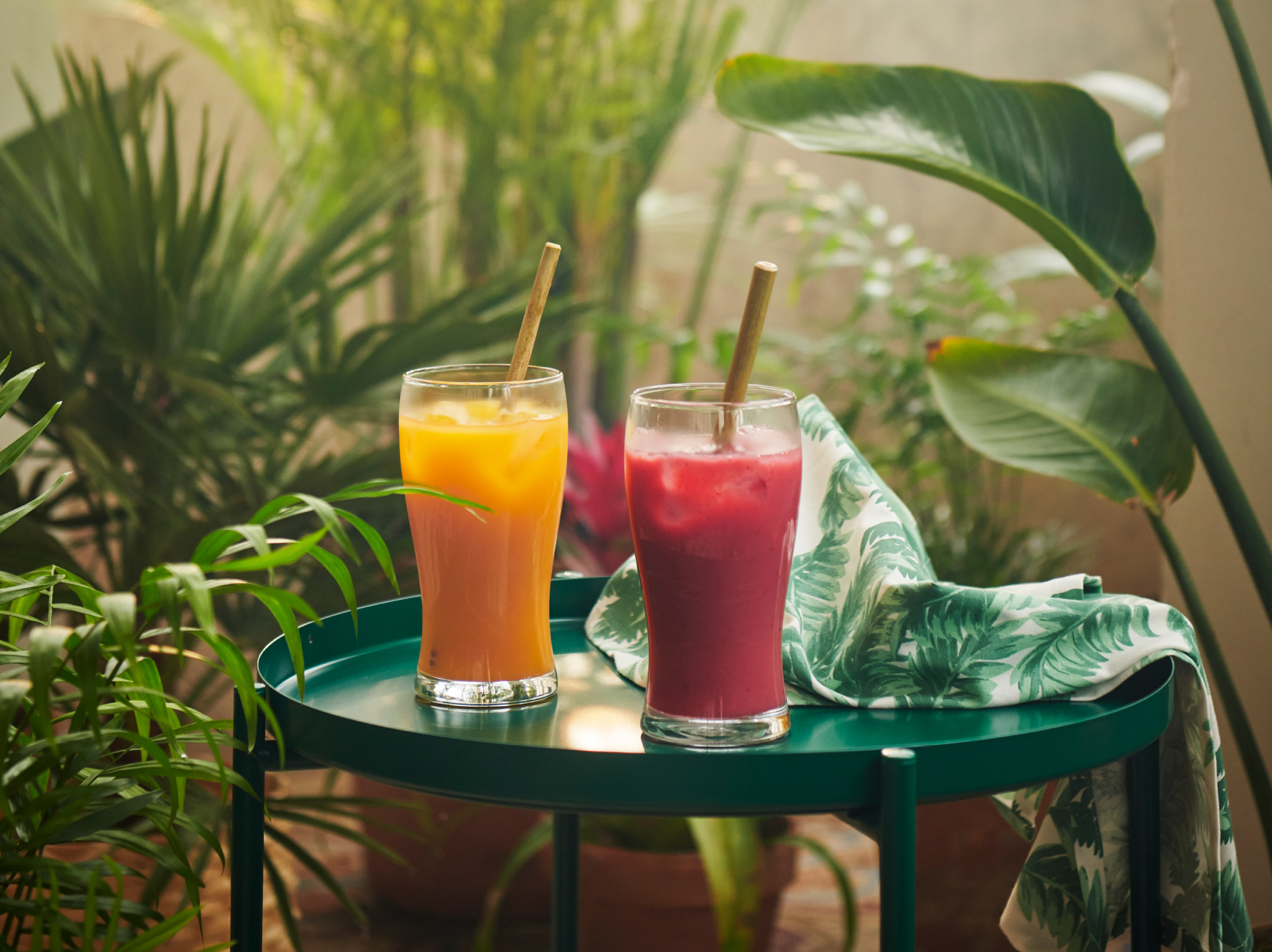 Two glasses with FRUKSTUND pre-blended smoothie mixes in tropical yellow and strawberry red placed on a table among plants.