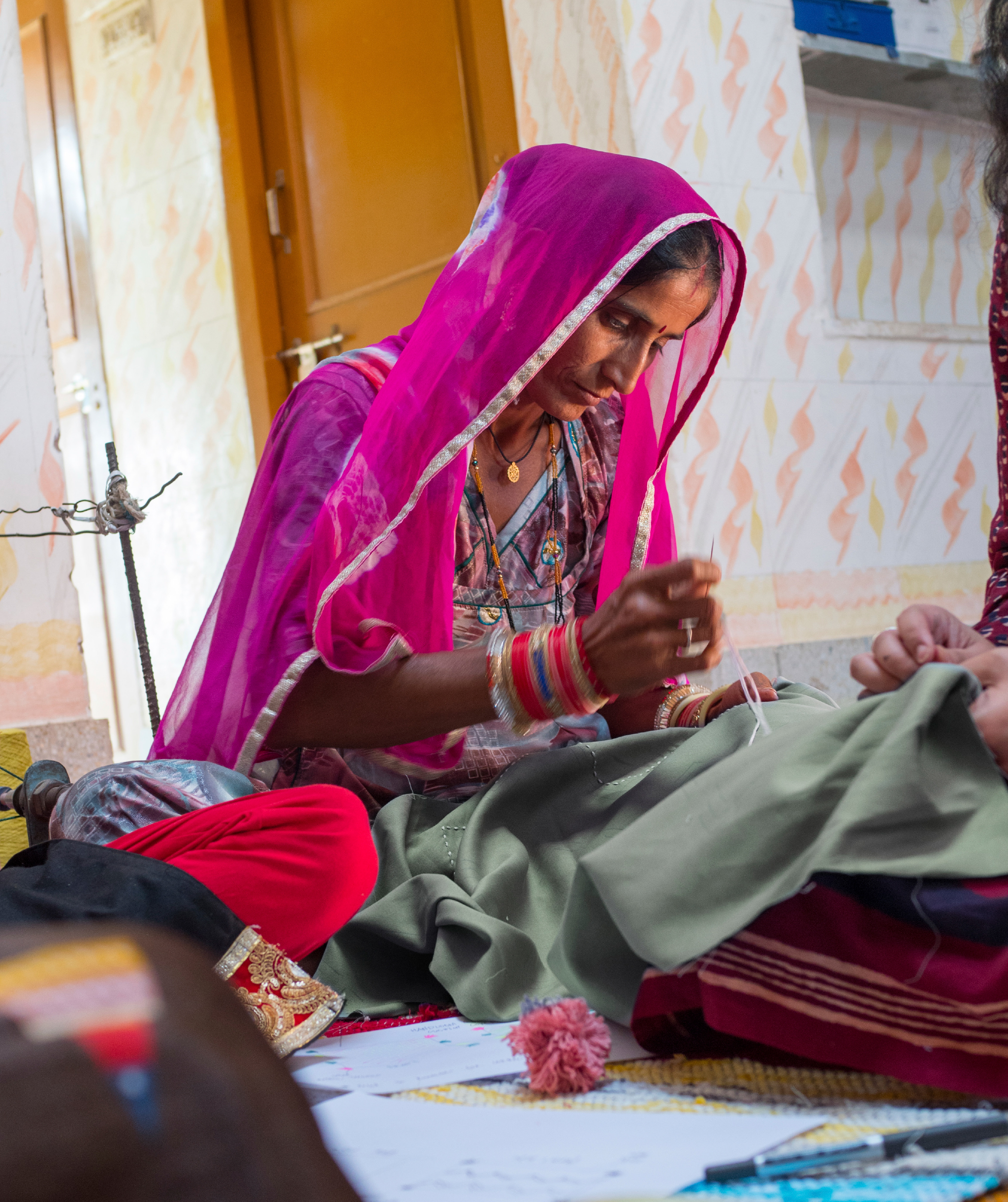 Woman dressed in a colourful dress and sheer shawl sitting on a rug with other women, doing needlework on a piece of fabric.