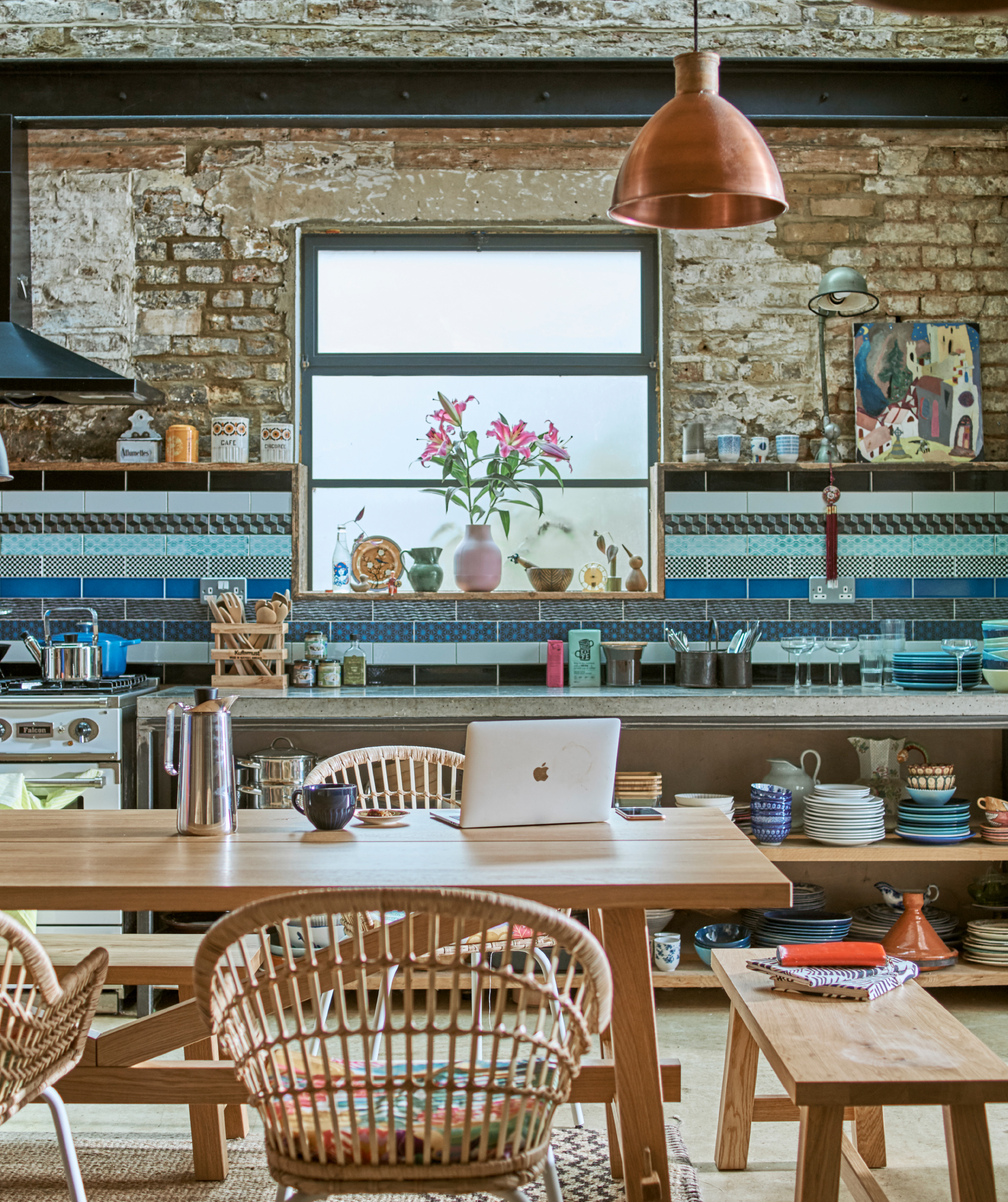 Wood table with benches and rattan chairs in an industrial kitchen with blue wall tiles and crockery piled on open shelves.